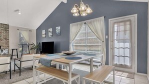 Un comedor moderno con techo alto, una araña y un amplio ventanal con persianas.
