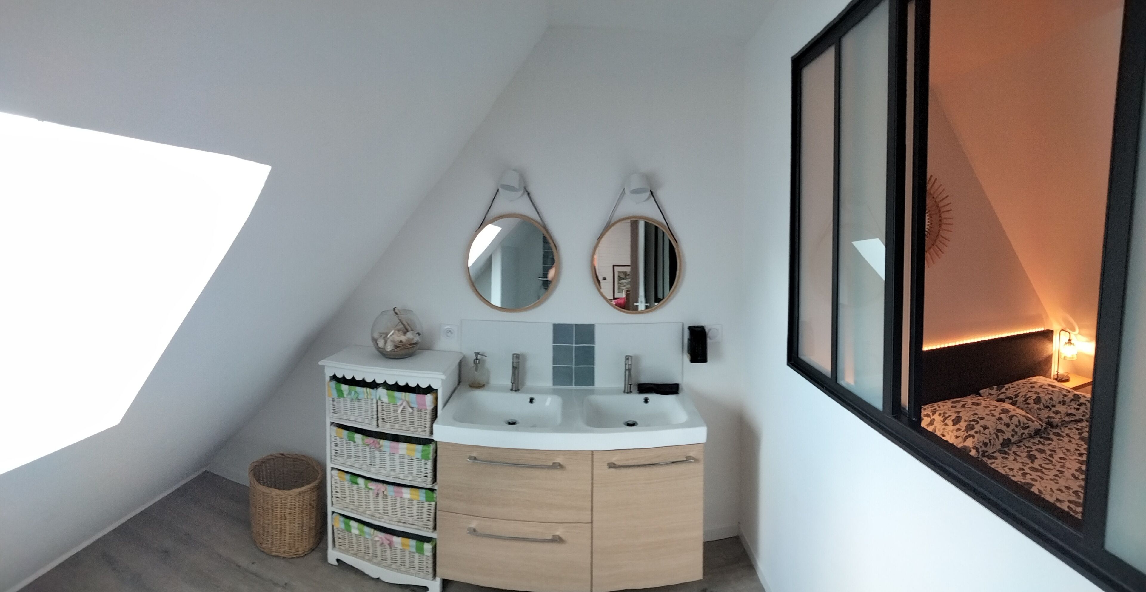 A modern bathroom with a skylight, a white sink, two mirrors, and a bed with a patterned bedspread.