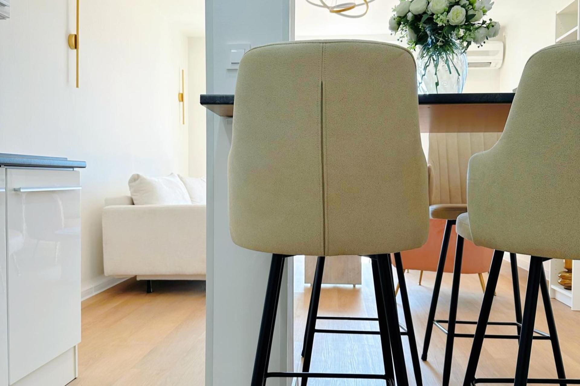 A modern kitchen with a white countertop, two bar stools, and a white sofa in the background.