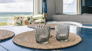 Un coin repas avec vue sur l’océan, comprenant une table agrémentée de sets de table en fibres tressées, deux verres et une lampe murale bleue.