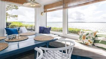 A sunlit room with a view of the water, featuring a blue table, white chairs, and a bench with cushions.