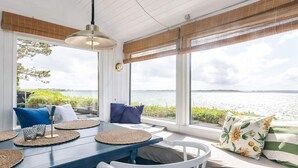 Une pièce baignée de lumière naturelle, avec vue sur l’eau, comprenant une table bleue, des chaises blanches et un banc agrémenté de coussins.