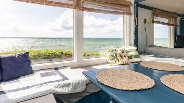 Une pièce avec vue sur l’océan, une table bleue avec des sets de table en fibres tressées, un banc rembourré et un canapé avec des coussins.