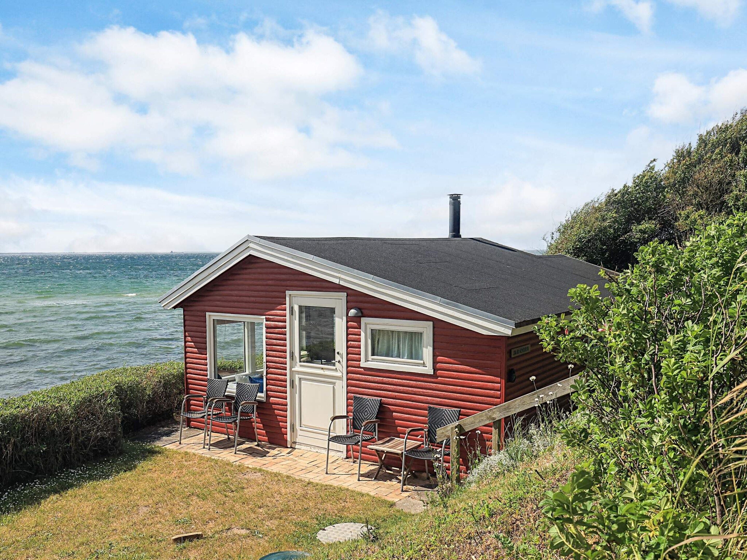 A red log cabin with a porch, a chimney, and a view of the sea.
