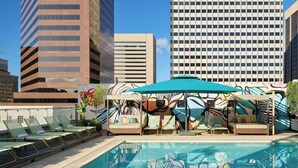Un área de piscina en la azotea con sillones de descanso y una pérgola, rodeada de edificios altos.