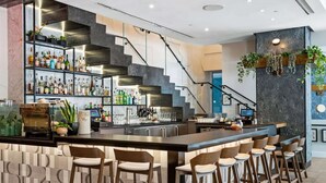 Zona de bar moderna con mostrador, varias estanterías llenas de botellas de licor, una máquina de café y taburetes.