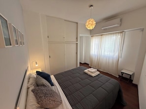 A hotel room with a large bed, a bedside lamp, a wardrobe, a window with curtains, and an air conditioning unit.