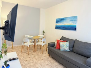 A living room with a grey sofa, a painting on the wall, a TV on a stand, a dining table with chairs, and a small plant on a table.