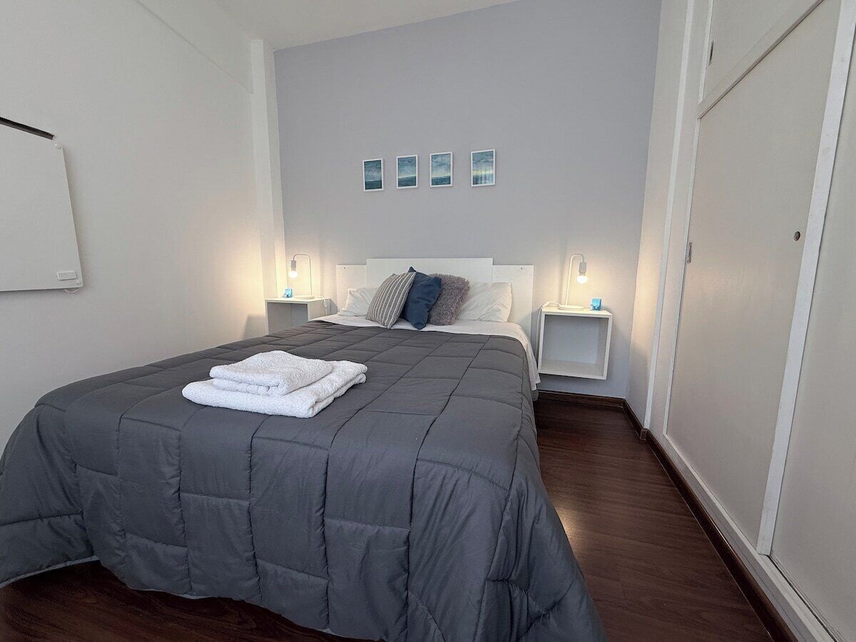 A neatly made bed with a grey duvet, a white towel, and a headboard with pillows. There are two bedside tables with lamps, and a wall-mounted shelf above the bed.