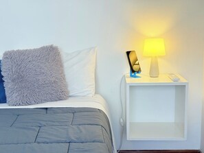 Ein Bett mit grauer Daunendecke und einem flauschigen Kissen, ein weißer Nachttisch mit einer Lampe und ein Smartphone darauf.