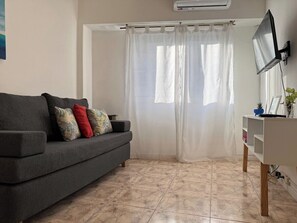 A living room with a grey sofa, a white curtain, a flat-screen TV, and a tiled floor.