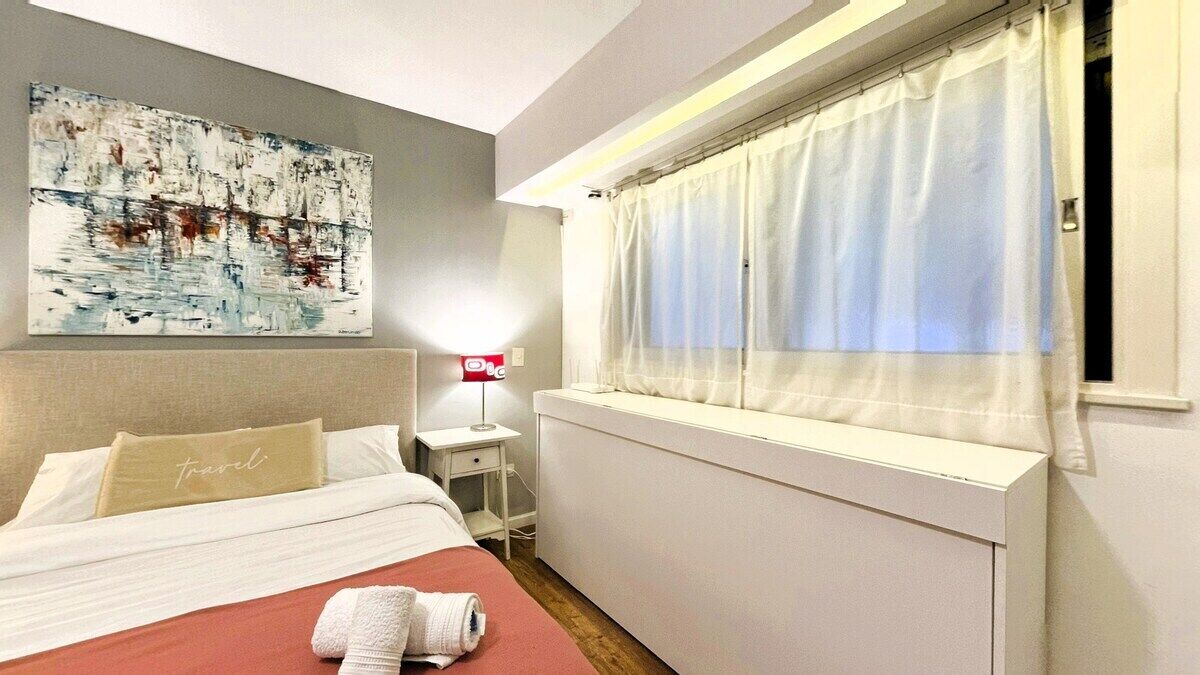 A hotel room with a bed, a bedside table, a lamp, a window with curtains, and a painting on the wall.