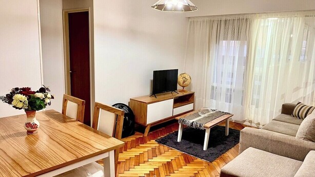 A living room with a wooden dining table, a sofa, a TV stand with a globe, and a black rug.