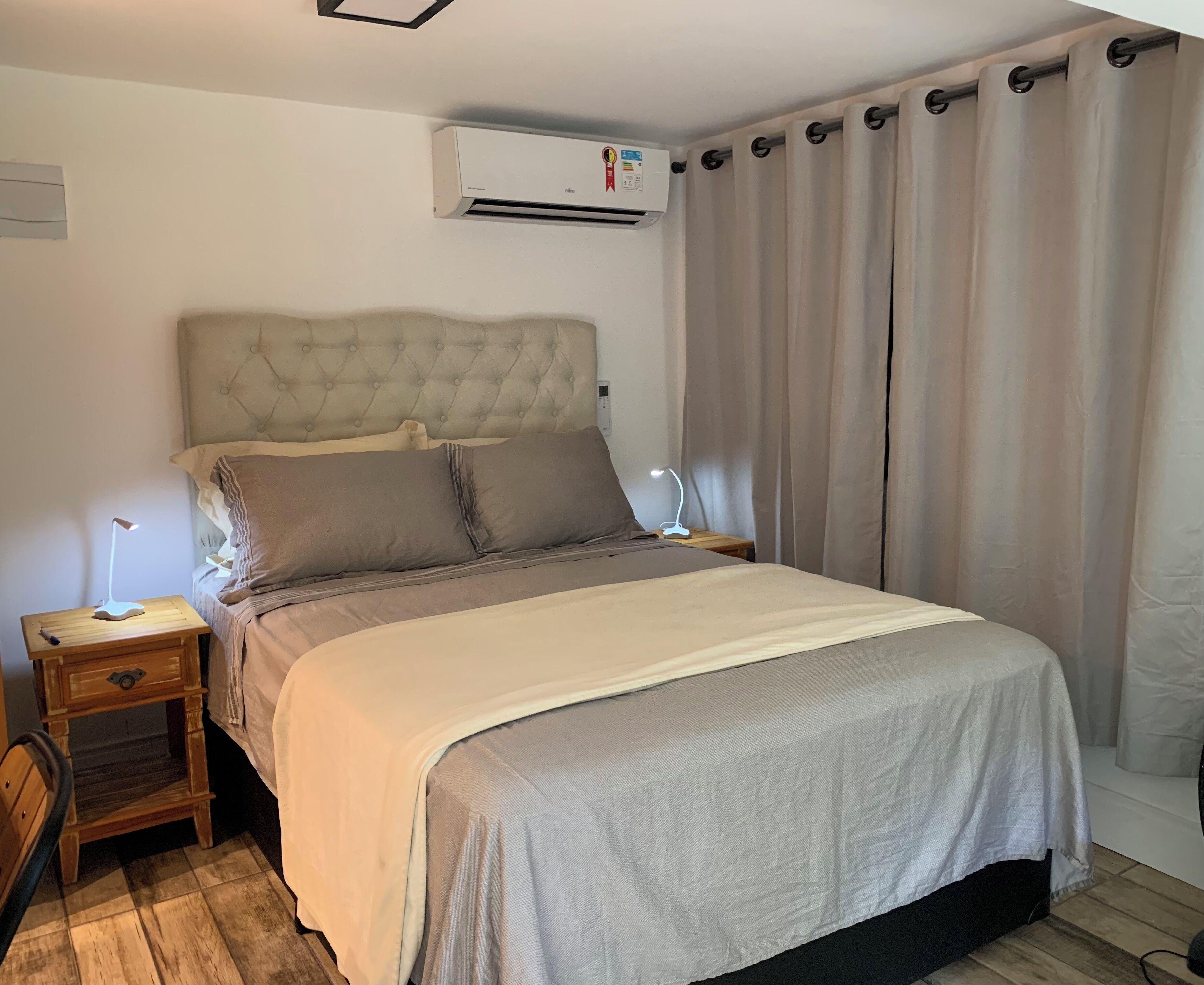 Een slaapkamer met een gestoffeerd hoofdeinde, een bed met grijze beddengoed, een nachtkastje met een lamp, en een wandairconditioner.
