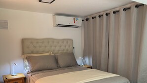 A bedroom with a tufted headboard, a bed with grey bedding, a bedside table with a lamp, a nightstand, and a wall-mounted air conditioner. - House - Studio apartment for couples in a gated community with pool, Praia do Curral in Ilhabela. (Ribeirão Ilhabela)