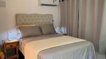 Een slaapkamer met een gestoffeerd hoofdeinde, een bed met grijze beddengoed, een nachtkastje met een lamp, en een wandairconditioner.
