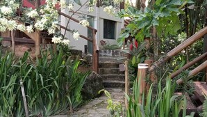 A stone pathway leading to a wooden staircase surrounded by lush greenery and flowering plants. - House - Studio apartment for couples in a gated community with pool, Praia do Curral in Ilhabela. (Ribeirão Ilhabela)