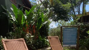A patio area with wicker lounge chairs and a small round table, surrounded by lush tropical plants and a wooden deck. - House - Studio apartment for couples in a gated community with pool, Praia do Curral in Ilhabela. (Ribeirão Ilhabela)