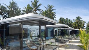 A modern, glass-walled mobile home with a curved design, situated in a tropical setting with palm trees in the background. - Sunny Pavilion (Thoddoo)