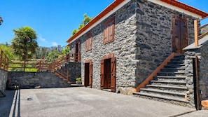 Ein Steinhaus mit einer Treppe, die zu einer Holztür führt, umgeben von einer gepflasterten Fläche und einer Steinsäule.