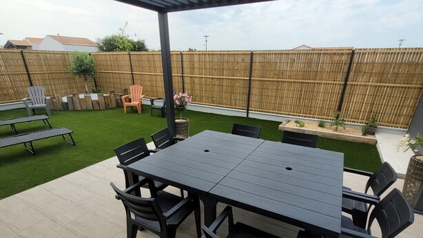 A covered patio with a table and chairs, a wooden fence, and a grassy area in the background. - New house Porte de Noirmoutier (Beauvoir-sur-Mer)