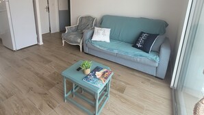A living room with a light blue sofa, a small table with a plant and a book, a white refrigerator, and a wall with a sign. - New house Porte de Noirmoutier (Beauvoir-sur-Mer)