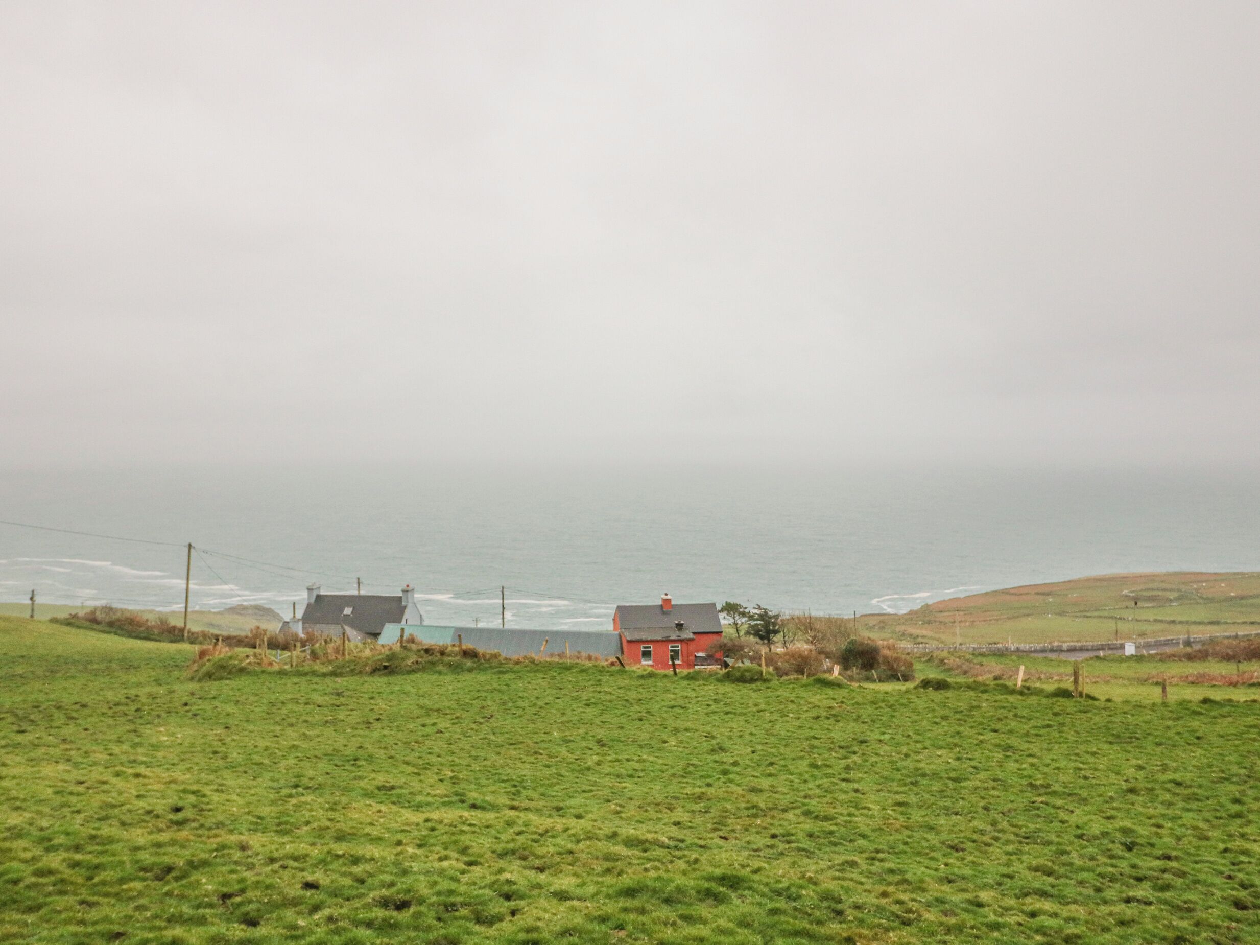 Paisaje costero con una casa roja y dos edificios más al fondo, rodeado de campos verdes y vistas al mar.