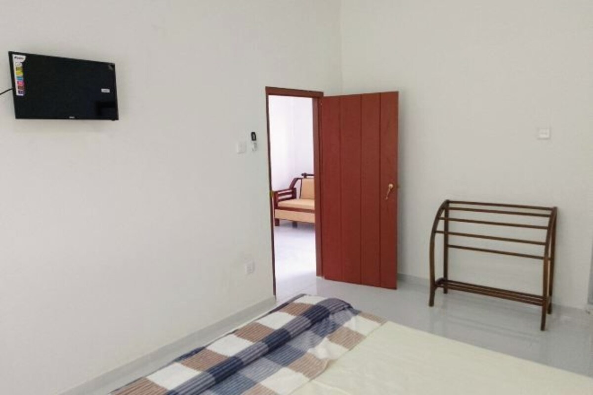 A room with a bed, a wooden door, a metal bench, and a wall-mounted TV.
