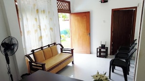A living room with a sofa, a table, a fan, a small table with a device, and a door leading outside. - Tropical Suites (Negombo)