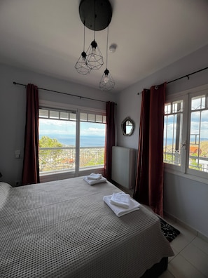 A bedroom with a large bed, two hanging pendant lights, a view of the sea, and a window with curtains.