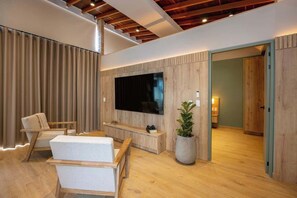 Ein modernes Wohnzimmer mit einem großen Flachbildfernseher an einer Holzwand, einer Sitzecke mit Sofa und Sesseln, einer Topfpflanze und Holzboden.