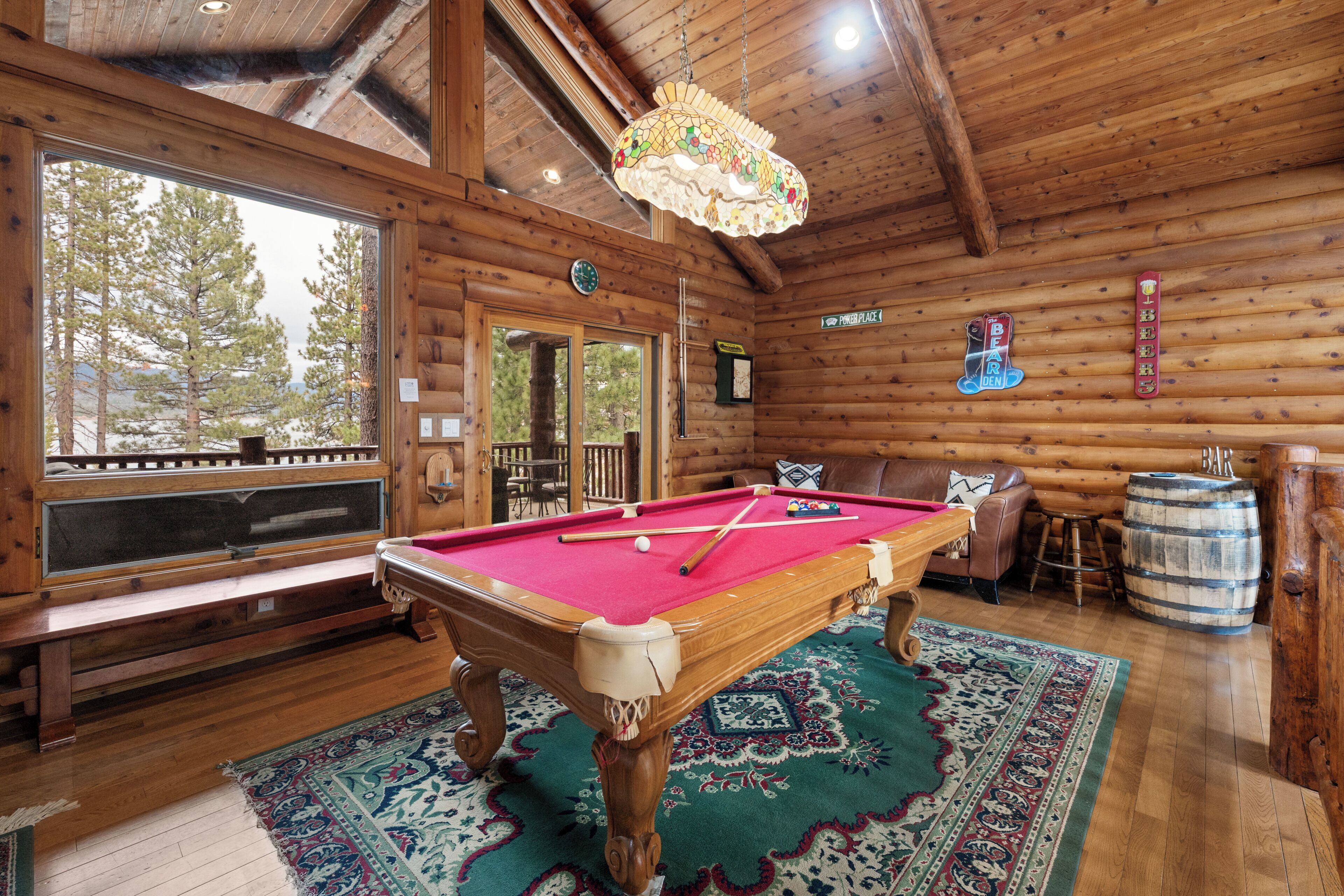 A wooden cabin interior with a pool table, a bench, a bar, and a large window overlooking trees.