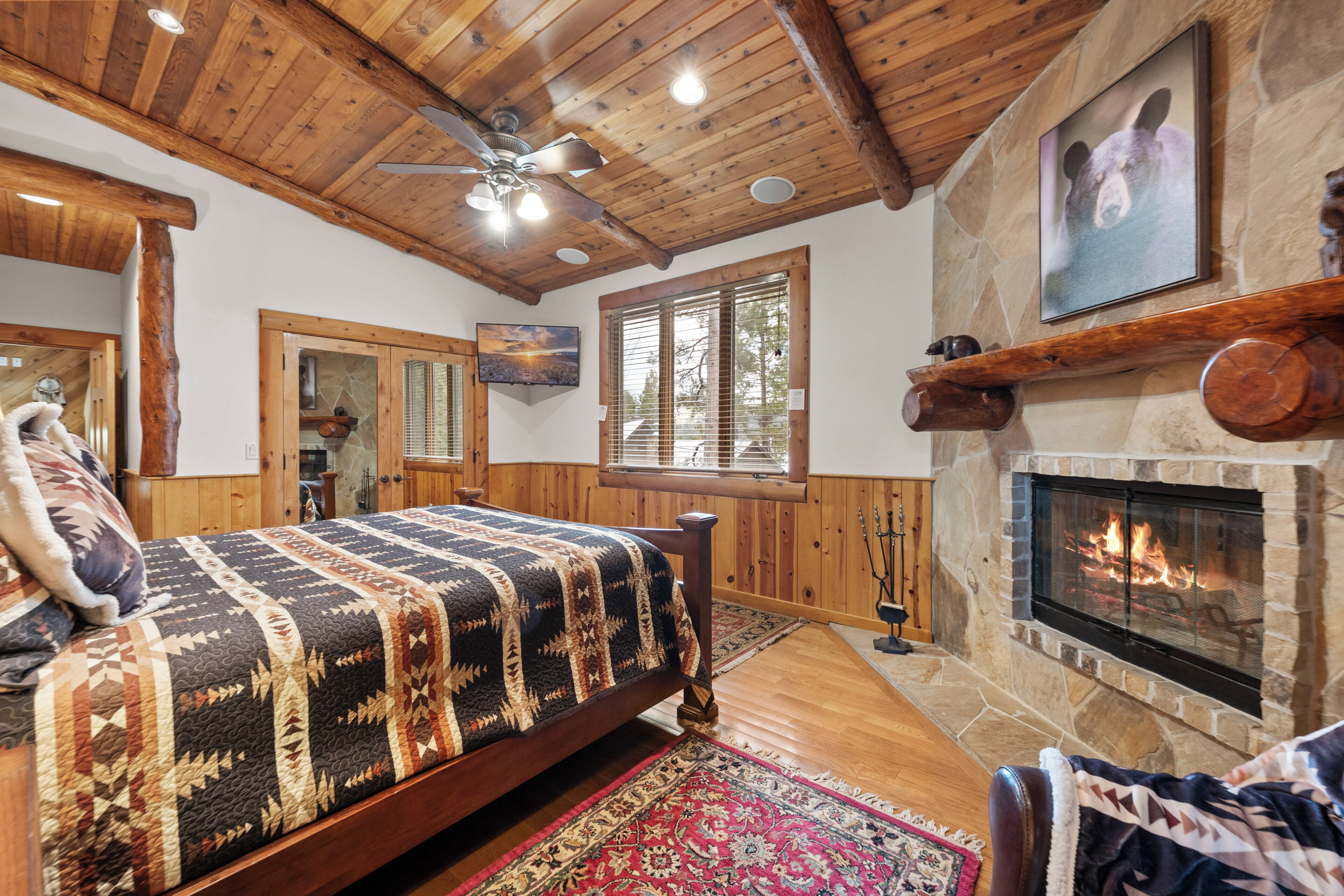 A cozy room with a wooden ceiling, a fireplace, a bed with a patterned quilt, and a large window.