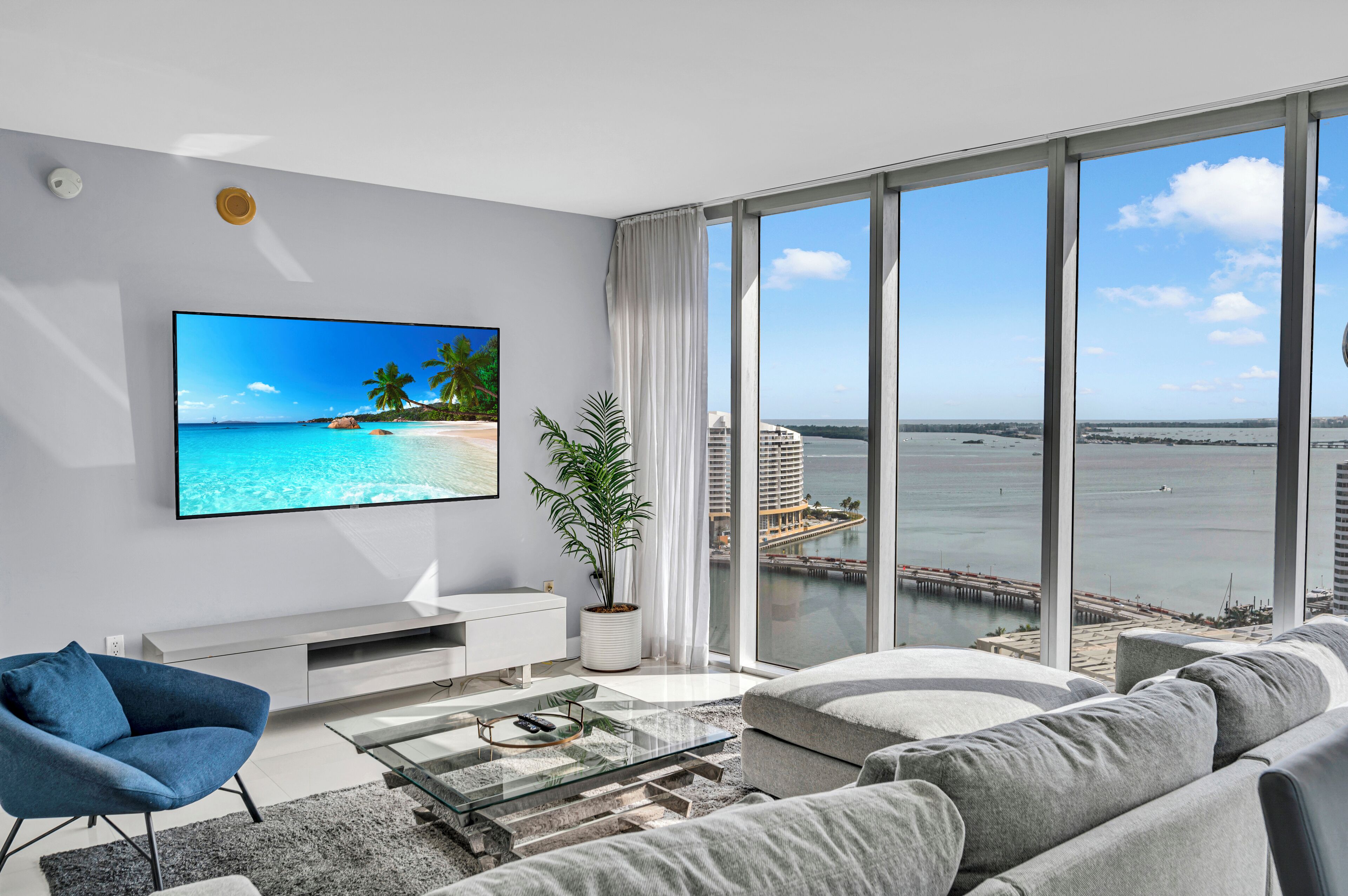 A modern living room with a large TV displaying a beach scene, a sofa, a coffee table, and a view of the ocean through large windows.