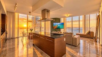A modern living room with a large TV, a dining area with a glass table, and a kitchen with a central island.