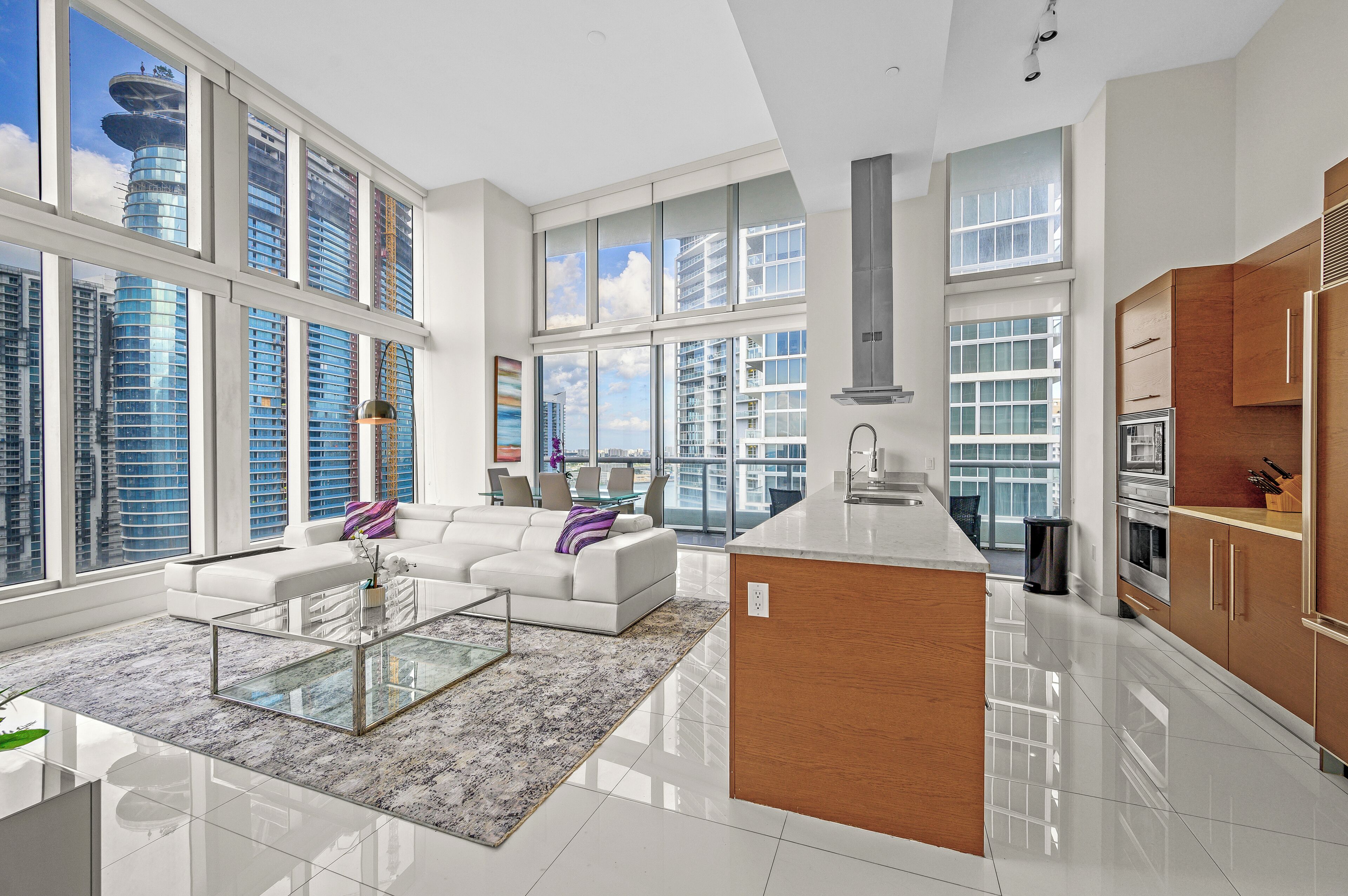 A modern apartment with large windows, a living area with a sofa and coffee table, and a kitchen with wooden cabinets and stainless steel appliances.