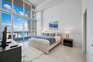 Un dormitorio moderno con un ventanal grande, una cama con manta gris, dos lámparas de noche y un cuadro en la pared.