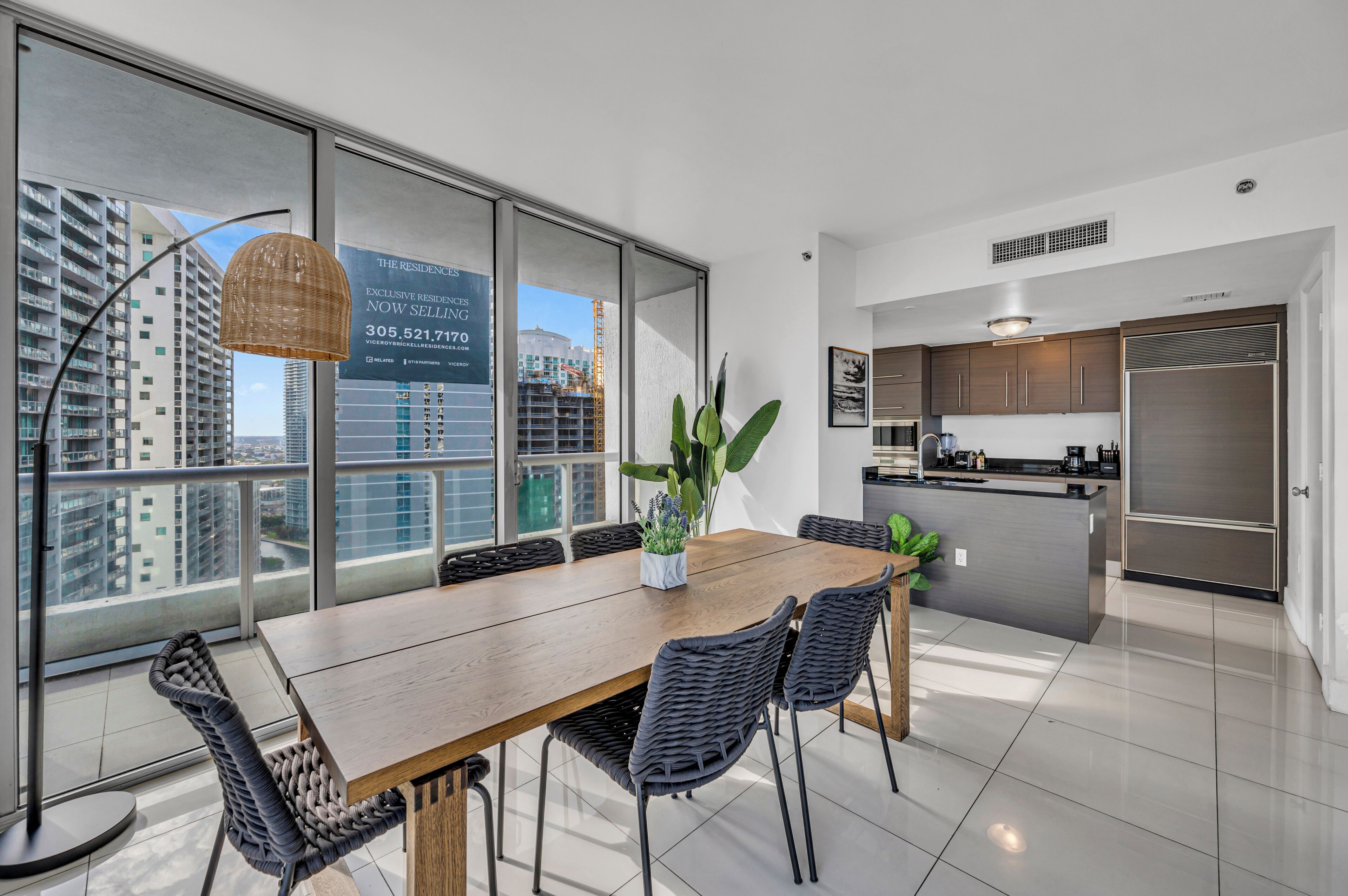 A modern kitchen with stainless steel appliances, a dining area with a wooden table and wicker chairs, and large windows offering city views.