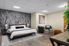 A modern bedroom with a large bed, a desk, and a chair. There is a wall with a tree branch pattern and a painting on the wall.