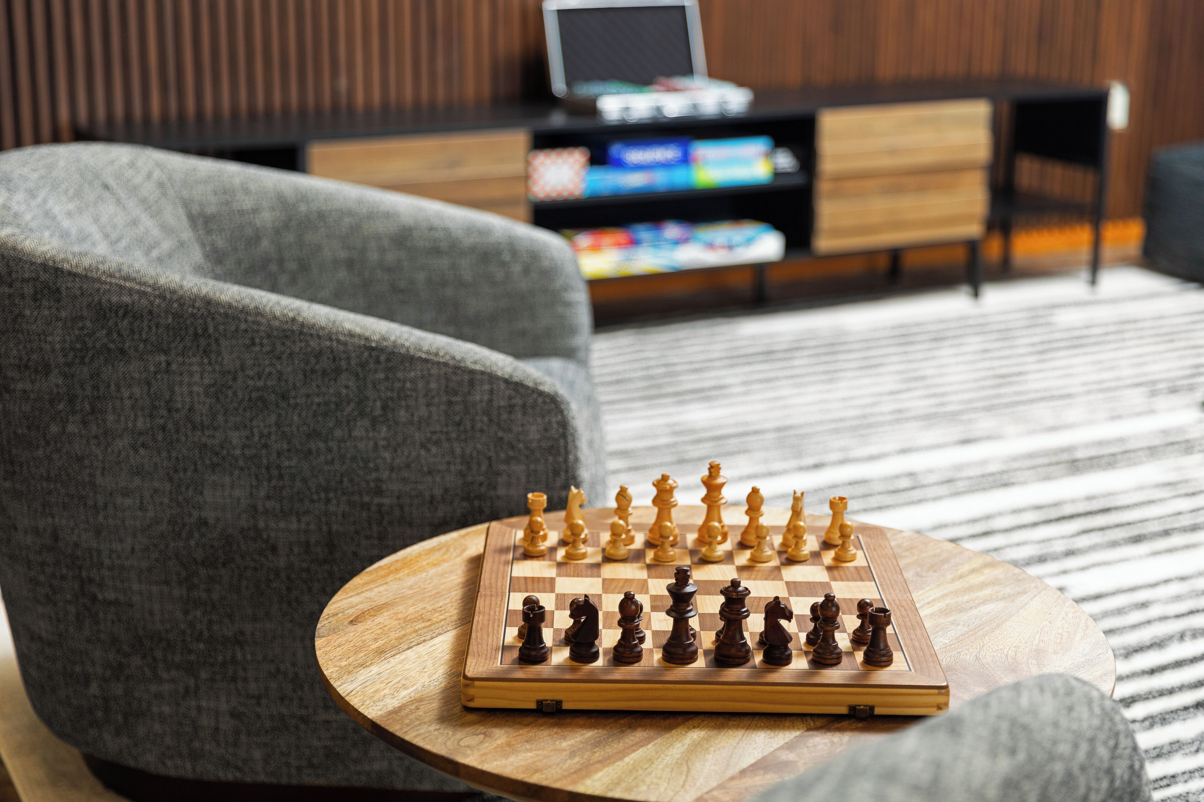 A chessboard on a round wooden table in a modern living room with a grey sofa and a wooden cabinet in the background.