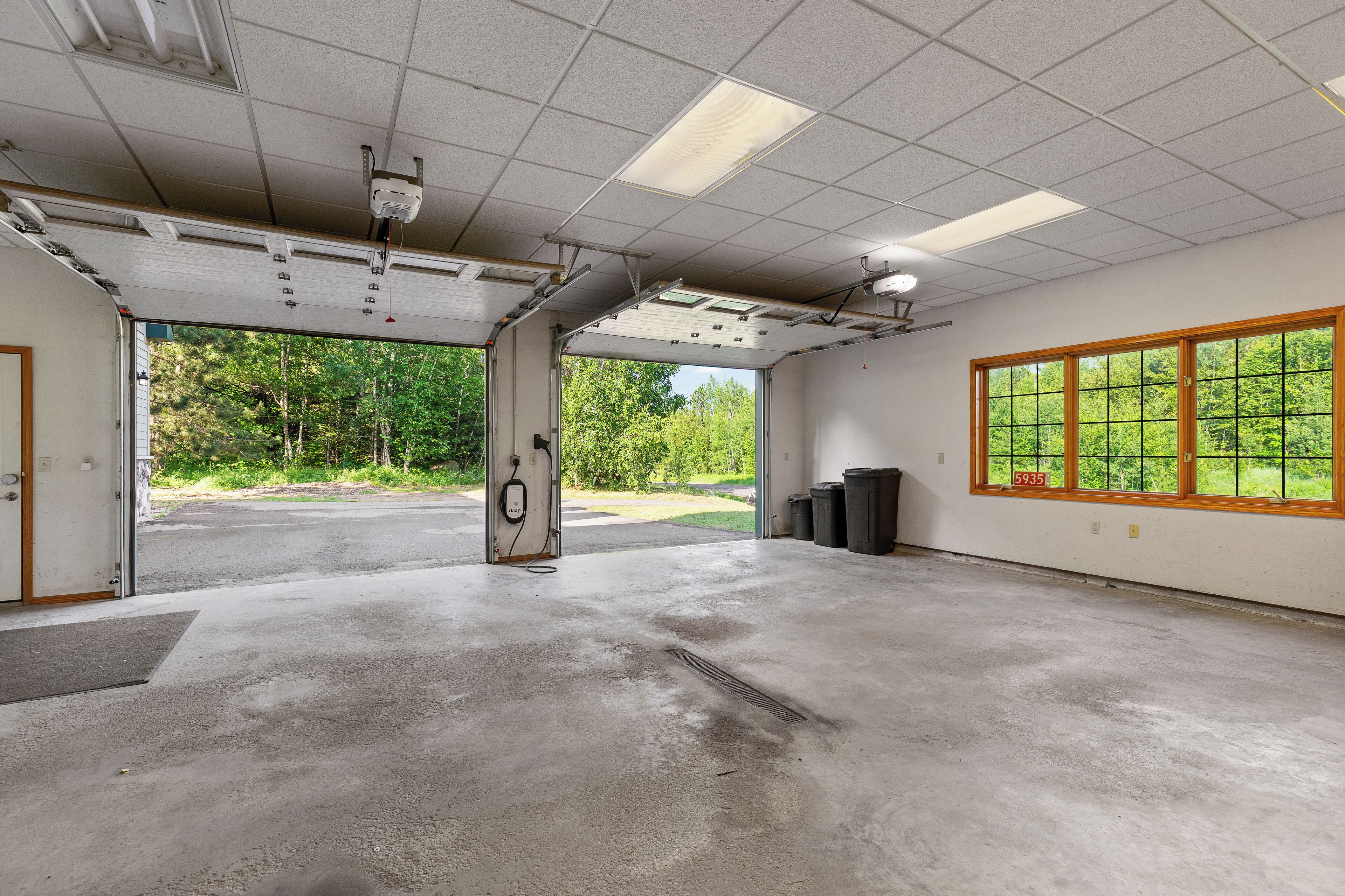 A spacious indoor area with a concrete floor, a large window with a view of greenery, and a ceiling with fluorescent lights and a smoke detector.