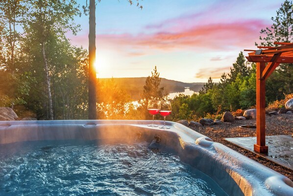 A hot tub with two glasses of red wine, surrounded by trees and a wooden pergola, with a sunset in the background.