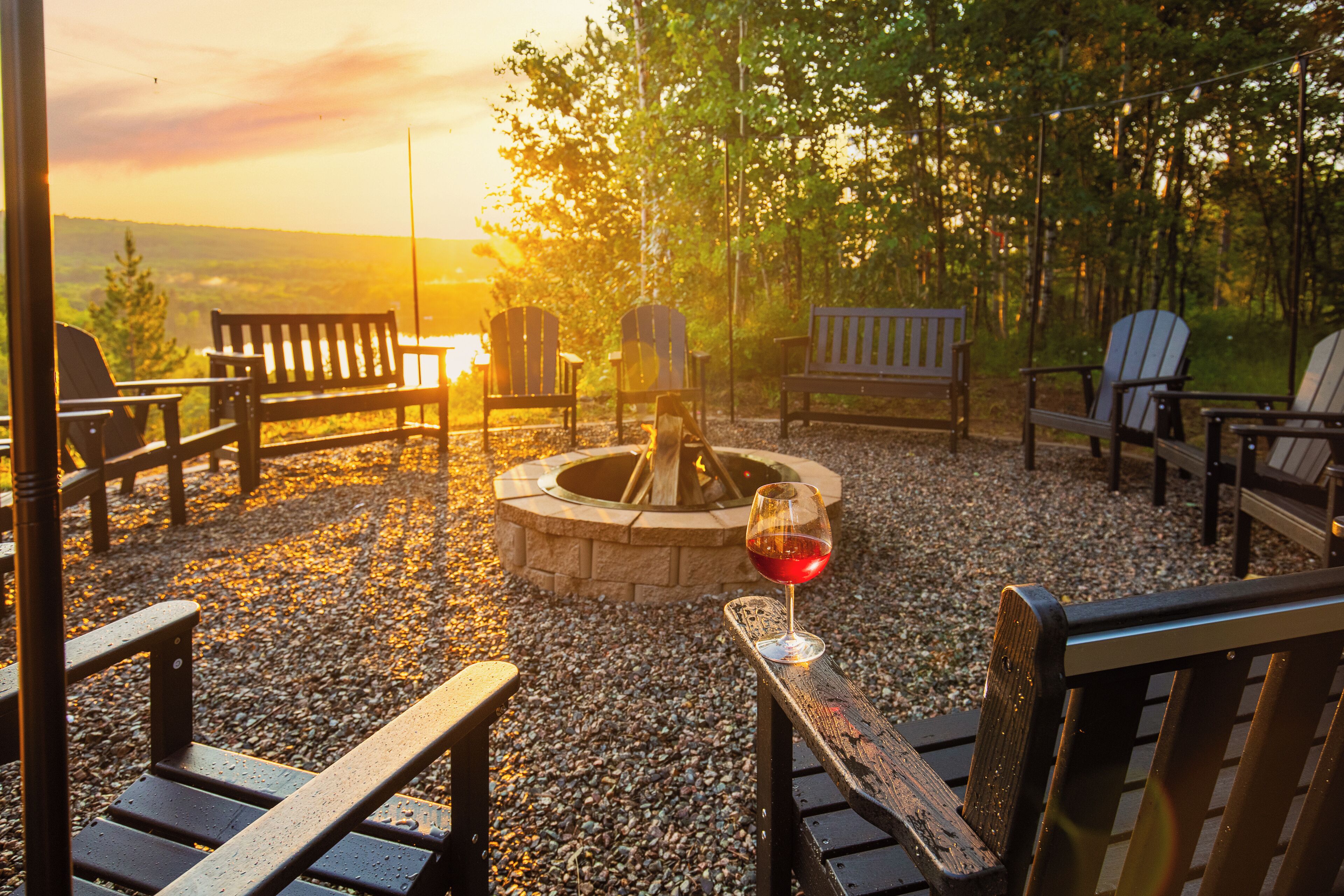 A fire pit surrounded by wooden benches and chairs in a gravel area with trees in the background.