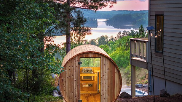 En cylindrisk träkonstruktion med en dörr och fönster, belägen i ett parkliknande område med utsikt över en sjö och kullar.