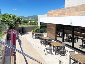 An outdoor patio area with multiple tables and chairs, a glass railing, and a view of the surrounding landscape.