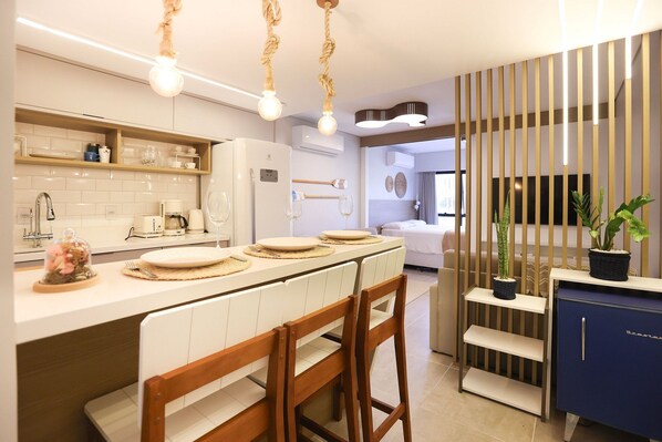 A modern kitchen with white cabinetry, a built-in oven, and a coffee machine. There are wooden chairs and a blue cabinet with a handle.
