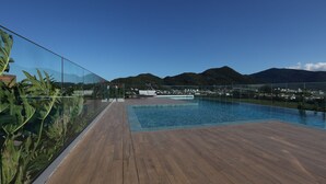 Terraza de madera con piscina y barandilla de vidrio con vistas a una zona residencial y montañas.