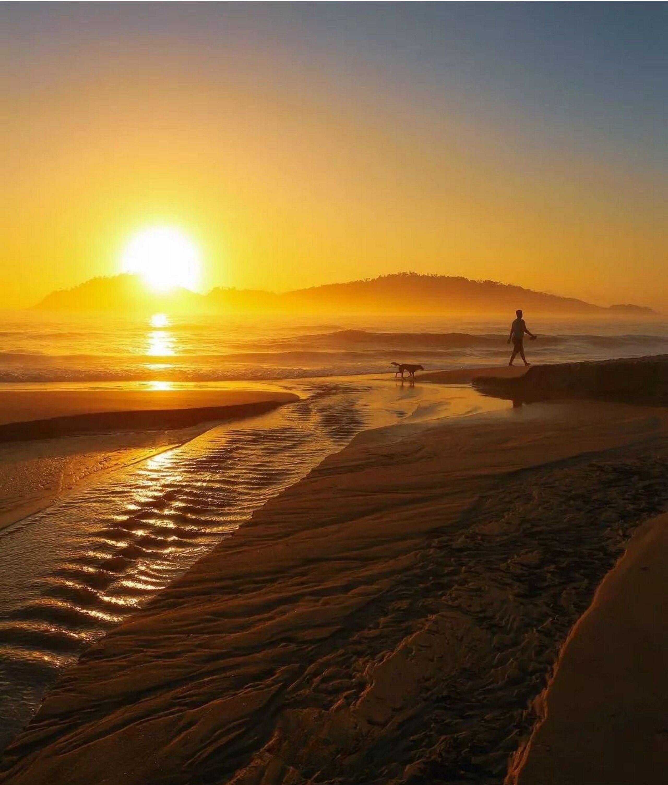 A beach scene at sunset with a person and a dog walking along the shore.