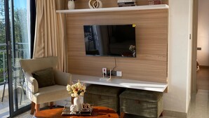 Ein modernes Wohnzimmer mit einem an einer Holzwand befestigten Flachbildfernseher, einem bequemen Sessel, einem Couchtisch mit Blumenvase und einem großen Fenster mit Vorhängen.