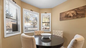 A dining area with a round table, four chairs, and a bowl in the center. There are three windows with blinds, and a bear painting on the wall.